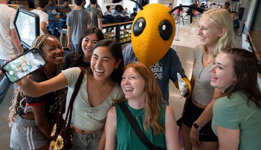 A group of smiling Georgia Tech undergraduate students take a photo with Buzz, Tech’s yellow jacket mascot. In the background, the interior of the student center is visible.
